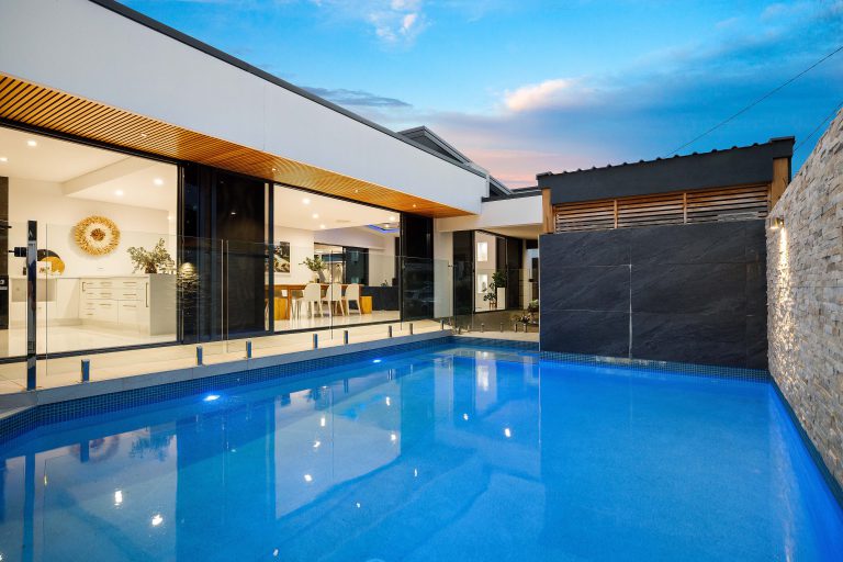 Saltwater pool at twilight with stone feature wall, glass fencing and illuminated kitchen visible through full-height glass doors at renovated Southport home