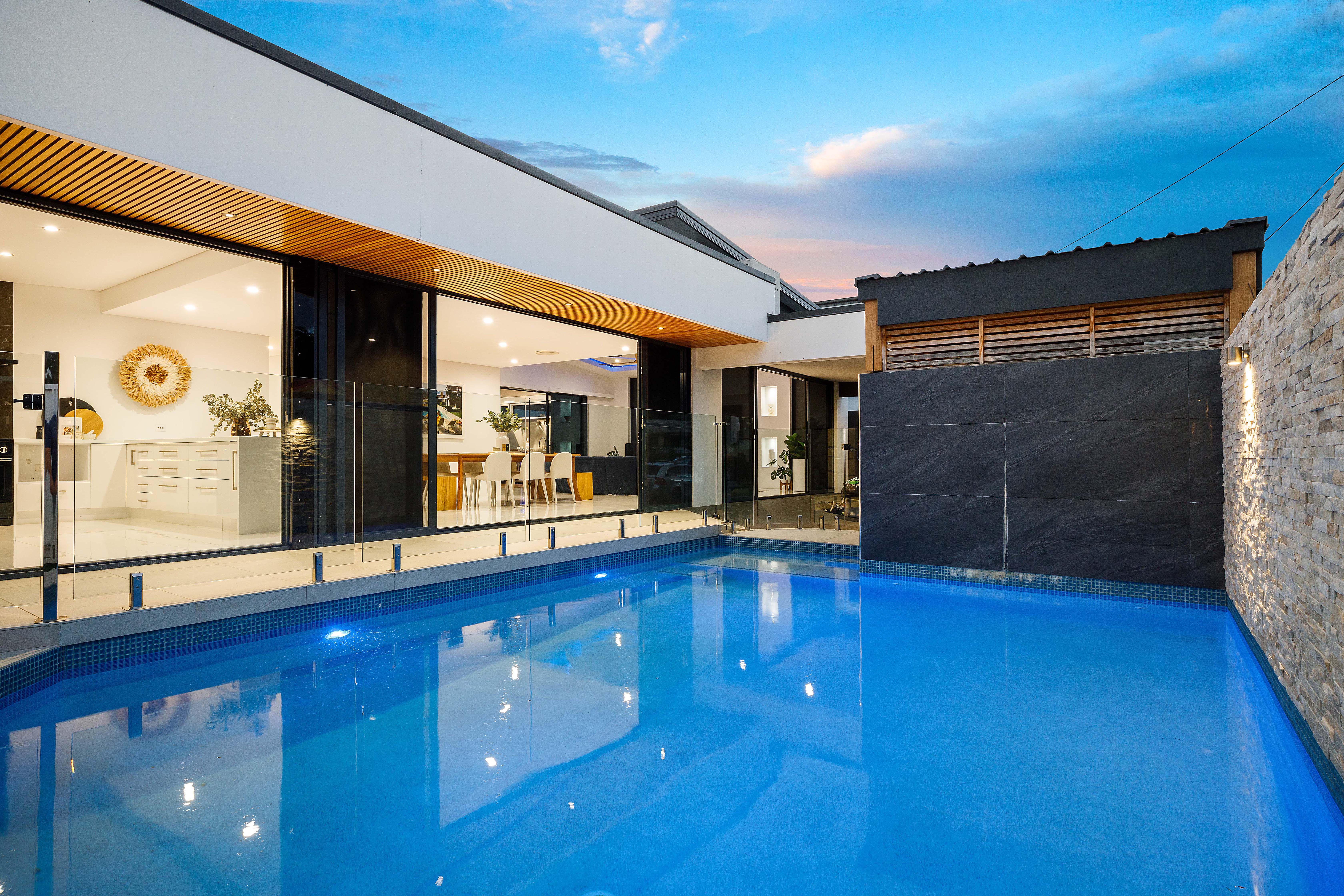 Saltwater pool at twilight with stone feature wall, glass fencing and illuminated kitchen visible through full-height glass doors at renovated Southport home