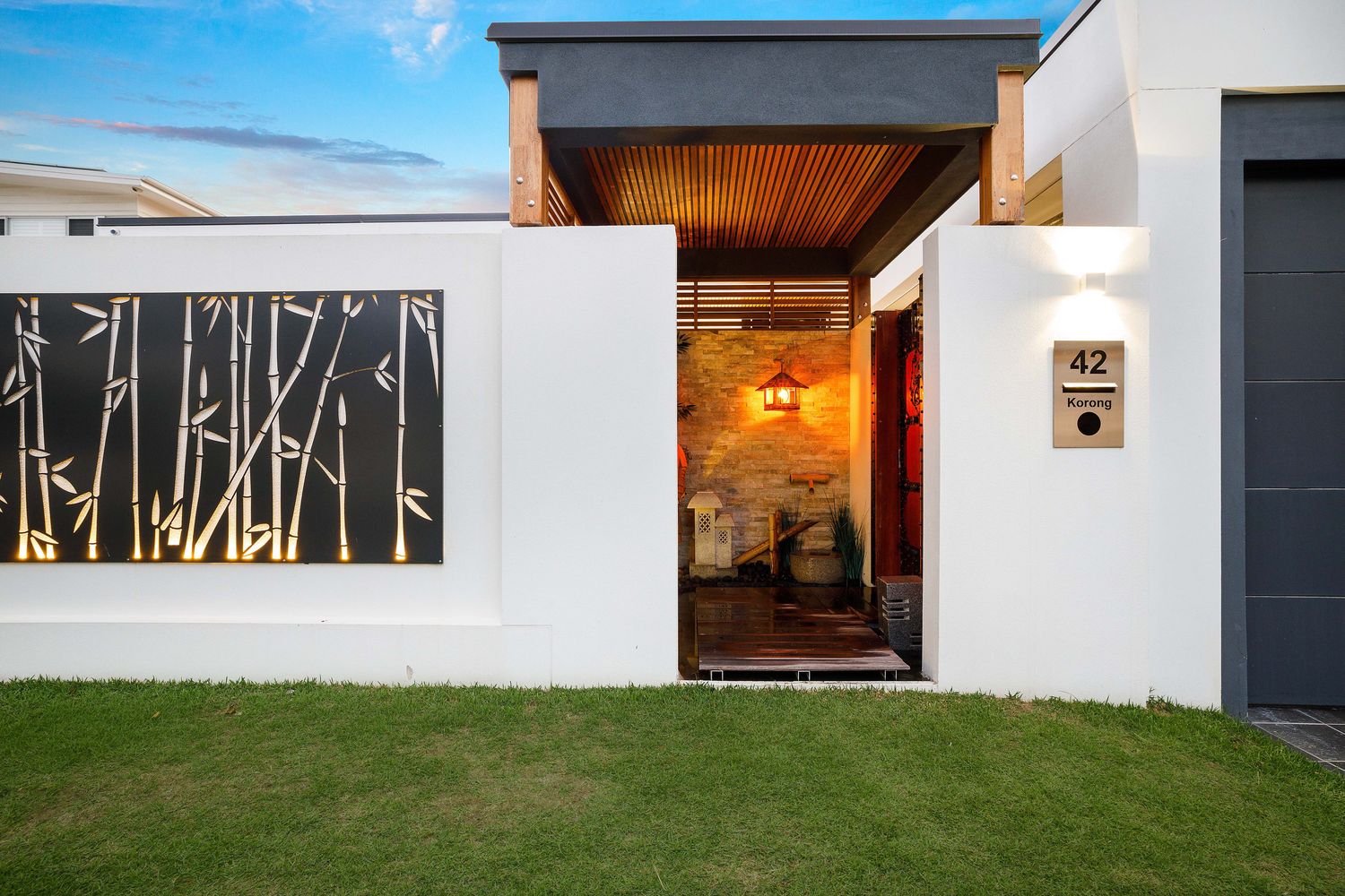 Modern front facade at dusk with laser-cut bamboo screen, timber batten soffit, sandstone feature wall and manicured lawn at renovated Southport home